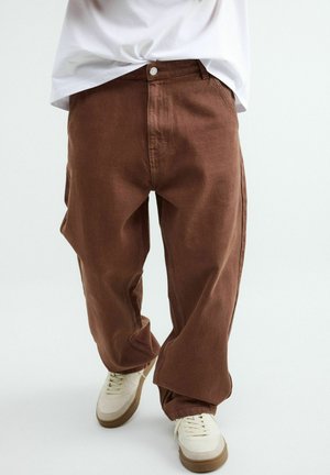 Osoba ubrana w luźne brązowe spodnie i białe sneakersy, idąca na jednolitym jasnym tle.