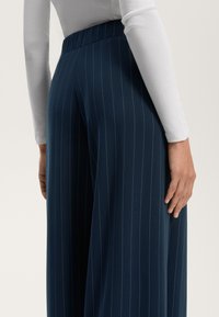 Temno modri hlače z visokim pasom in vertikalnimi črtami, izdelane iz gladkega blaga; prikazane s hrbtne strani z belo majico dolgimi rokavi.