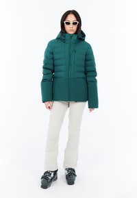 Puffy bunda v tyrkysové barvě s vysokým límcem a zipem v přední části, obsahující velké prošívané panely a boční kapsy. Nosit s béžovými kalhotami a černými lyžařskými botami.
