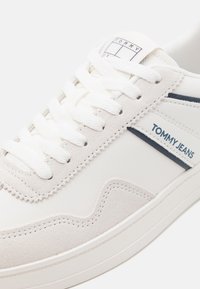 Bijela tenisica koja sadrži kožu i velur, sa vezicama, teksturiranom potplatom i "TOMMY JEANS" oznakom u tamnoplavoj boji.