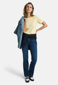 Lichtgele t-shirt, blauwe denimjack over de arm, donkerblauwe jeans en marineblauwe blokhakken, minimale branding op het bovenstuk.