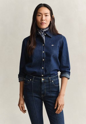 Vrouw draagt een donkere denim blouse met opgerolde mouwen, donkere denim jeans en een gestreepte sjaal om de hals, staand tegen een neutrale achtergrond.
