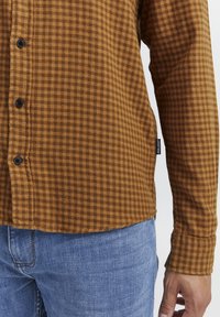 Camisa de franela de cuadro marrón con botones negros y dobladillo curvado, combinada con jeans de mezclilla azules; se puede ver la textura de la tela.