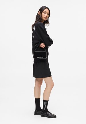 Sacoche noire en bandoulière, de forme rectangulaire, avec fermeture éclair et logo discret, associée à un sweat-shirt noir oversize et une jupe.