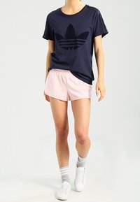 Marineblauwe T-shirt met een groot zwart logo, gecombineerd met lichtroze shorts. Model draagt witte sneakers en grijs gestreepte sokken. Eenvoudig ontwerp.