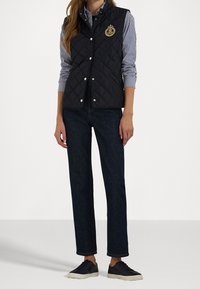 Marinblå quiltad väst med knappar framtill, logotypmärke, bärs över en blå- och vitrandig skjorta. Matchas med mörka jeans och svarta sneakers.