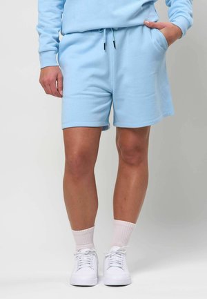 Személy világoskék rövidnadrágot és hozzá illő pulóvert visel, fehér tornacipőben és világos rózsaszín zokniban, egyik kezét a zsebébe dugva áll.