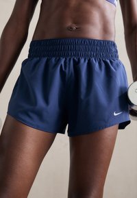 Persona atletica che indossa pantaloni corti Nike blu scuro e reggiseno sportivo, che tiene un manubrio, mostrando un addome e gambe tonici.
