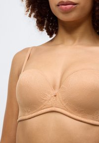 Čipkasti grudnjak u boji kože s dubokim izrezom, jastučićima, podesivim trakama i malim dekorativnim akcentom u sredini.