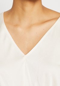 Krémový top s výstřihem do V, s hladkou texturou a jemnými švy. Tkanina vypadá lehce a prodyšně.