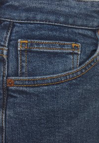 Närbild av en blå jeansficka med synliga gula sömmar och en liten kopparniting på vänster sida.