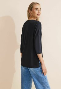 Schwarzes Oberteil mit 3/4-Ärmeln, weichem Stoff und lockerer Passform. Getragen mit hellblauen Jeans. Nahtdetails an den Schultern und Seiten sorgen für Textur.