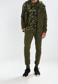 Grön camouflagehoodie och matchande byxor i sweatshirtmaterial; har dragkedja framtill, kenguruficka och ribbade muddar. Svarta sneakers.