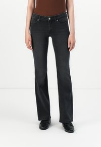 Slim-fit zwarte jeans gedragen met zwarte schoenen en een bruine mouwloze top, weergegeven op een rechtopstaande persoon tegen een effen witte achtergrond.