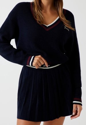 Femme portant un pull bleu marine à col en V avec des bordures rouges et blanches, assorti d'une jupe plissée coordonnée. « Guess » est inscrit sur le pull et la ceinture.