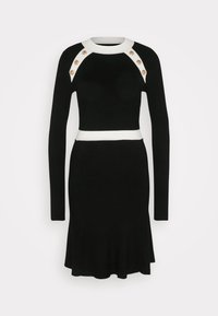 Robe en maille noire à manches longues avec bordure blanche au niveau de l'encolure et de la taille, ornée de six boutons dorés sur les panneaux blancs aux épaules.