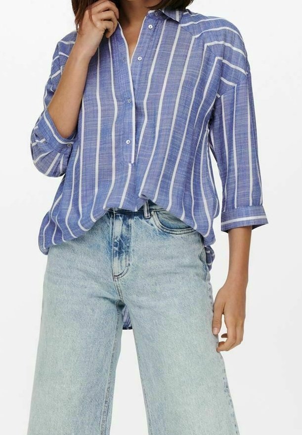 Camicia a righe blu e bianche con maniche arrotolate, realizzata in tessuto leggero. Abbinata a jeans a gamba larga in denim chiaro.