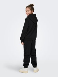 Melns džemperis ar priekšējo kabatu un regulējamu auklu, kopā ar atbilstošām sporta biksēm. Apģērbā ir ribota aproce un baltas sporta kurpes.