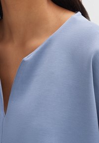 Top blu chiaro con scollatura a V. Il tessuto ha una texture liscia e morbida, con una vestibilità leggermente rilassata. Nessun motivo o accento visibile.