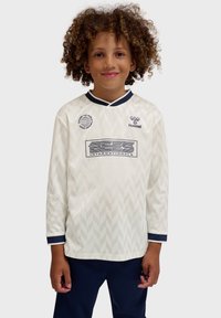 Enfant aux cheveux bouclés portant un maillot de sport blanc à motifs avec des logos et un pantalon bleu marine, debout devant un fond uni.