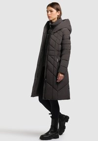 Nő áll egy világosszürke háttér előtt, hosszú fekete steppelt télikabátban, kapucnival, fekete leggings-ben és fekete bokacsizmában.
