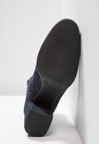Semelle noire d'une bottine en daim bleu marine avec talon bloc, taille 37, reposant contre un mur et une surface blanche.