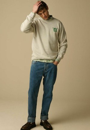 Young man in beige sweatshirt, green striped shirt, cuffed blue jeans, and brown shoes stands with one hand in pocket, the other touching his hair.