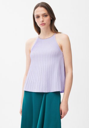 Junge Frau trägt ein ärmelloses, fliederfarbenes, geripptes Top mit Kettendetail und einen hoch taillierten türkisfarbenen Rock, steht vor weißem Hintergrund.