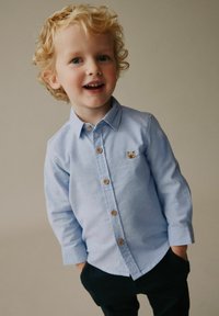 Cămașă albastru deschis cu nasturi, cu un mic emblemă de urs, nasturi din lemn, mâneci lungi și croială lejeră, asortată cu pantaloni închiși la culoare.