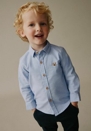 Svetlomodrá košeľa na gombíky s malým emblémom medveďa, drevené gombíky, dlhé rukávy a voľnejší strih, v kombinácii s tmavými nohavicami.