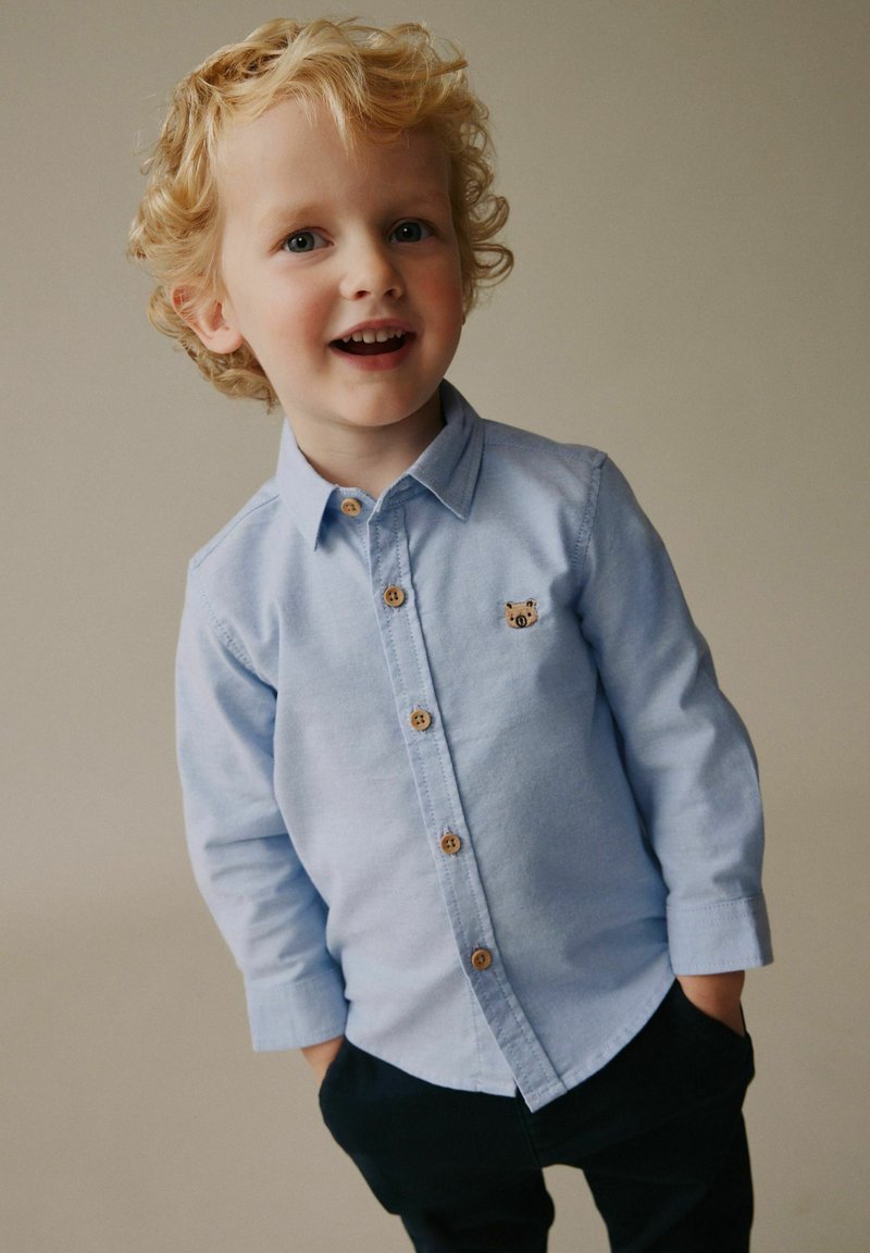 Cămașă albastru deschis cu nasturi, cu un mic emblemă de urs, nasturi din lemn, mâneci lungi și croială lejeră, asortată cu pantaloni închiși la culoare.