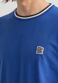 Niebieska bawełniana koszulka z okrągłym dekoltem, ozdobiona granatowo-białymi paskami oraz małym haftowanym logo tygrysa na piersi.