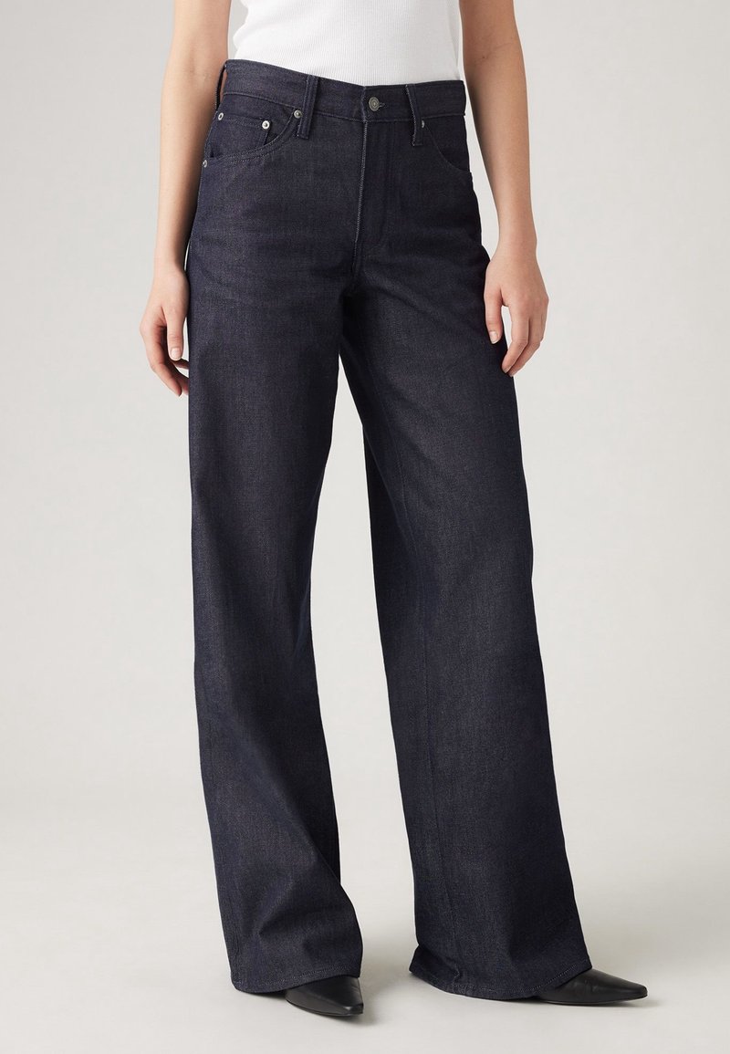 Personne portant un jean en denim sombre à jambes larges, associé à des chaussures noires à bouts pointus et un haut blanc, debout devant un fond uni.