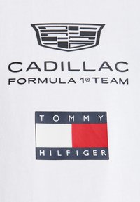 Λογότυπο Cadillac πάνω από το κείμενο "Cadillac Formula 1 Team" και το λογότυπο της σημαίας Tommy Hilfiger σε λευκό υφασμάτινο φόντο.