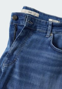 Jean en denim bleu plié avec poche avant visible, bouton, passant de ceinture et étiquettes intérieures indiquant la marque et la taille.