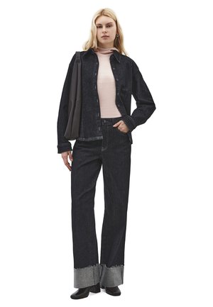 Femme portant une veste en denim noire et un jean à jambes larges avec de grands revers, un col roulé rose clair, des bottes noires et un sac à bandoulière noir.