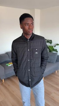 Un jeune homme portant une veste matelassée noire et un jean bleu clair se tient dans un salon avec un canapé gris et une plante verte en arrière-plan.