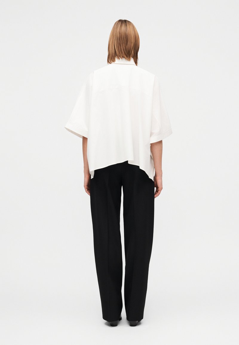 Chemise blanche oversize à manches courtes, ourlet asymétrique, associée à un pantalon large noir, les deux présentant des textures lisses.
