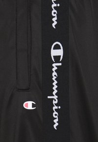 Černé sportovní šortky s texturovanou tkaninou, bílé logo "Champion" a malá vícebarevná nášivka na boku.