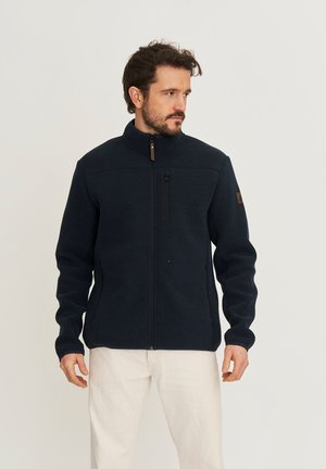 Man draagt een marineblauwe fleecevest met ritssluiting en witte broek, staand tegen een effen lichte achtergrond en kijkt naar zijn rechterkant.