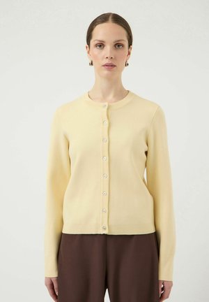 Femme à la peau claire et aux cheveux bruns attachés en chignon bas, portant un cardigan boutonné jaune pâle et un pantalon large marron foncé.