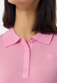 Ljusrosa polotröja med ribbad krage, fem matchande knappar och en liten logotyp på vänster sida. Mjuk textur.