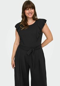 Svart overall med volangärmar, rund halsringning och knyt i midjan. Tyget verkar mjukt och lätt.