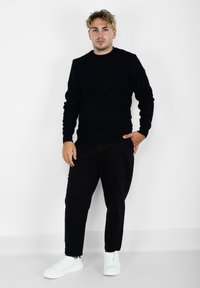 Maglione nero con collo rotondo, abbinato a pantaloni neri a gamba slim e sneakers bianche. Texture liscia, design semplice e senza motivi visibili.