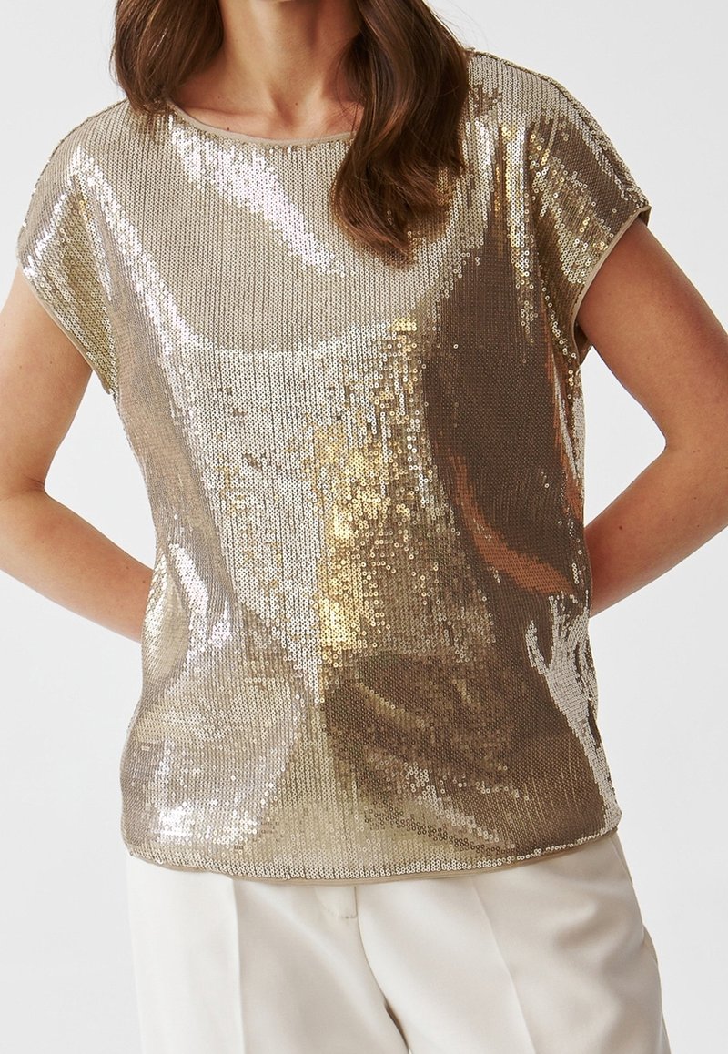 Femme portant un haut à manches courtes en sequins dorés brillants et un pantalon blanc, les bras pliés derrière son dos sur un fond uni.