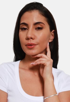 Weißes, geripptes Oberteil mit Rundhalsausschnitt, Model trägt ein silbernes Armband, lange dunkle Haare, glatte Haut, neutrale Lippenfarbe und dezentes Make-up.