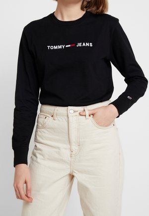 Kvinde iført sort, langærmet Tommy Jeans trøje stoppet ned i højtaljede cremefarvede jeans, med hånden i lommen, mod ensfarvet baggrund.