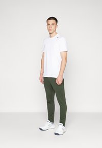 Biela krátka rukávová tričko, olivovo zelené slim-fit nohavice a biele športové topánky. Jednoduchý dizajn s logom na ramene; hladká textúra.