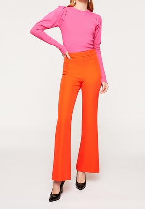 Femme portant un haut rose vif à manches longues avec des épaules bouffantes et un pantalon orange taille haute à jambes larges avec des talons hauts noirs.