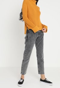 Personne portant un pull à col roulé moutarde, un jean gris délavé acide et des baskets noires, debout les mains près des poches.